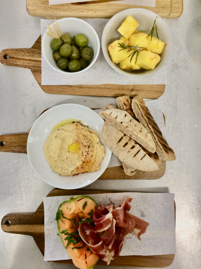 Een houten plank met olijven, polenta parmezaan, blokjes, hummus, brood, meloen en parmaham.